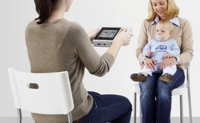 dwie kobiety siedzące na krześle, jedna trzyma urządzenie autorefraktometr Plusoptix, a druga dziecko