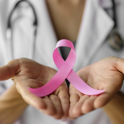 zdjęcie przedtawia na pierwszym planie obrócone dłonie, które trzymają różową wstążkę onkologiczną a w tle widać postać kobiety lekarza w białym kitlu i przewieszonym stetoskopem
