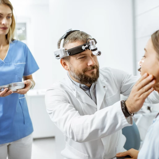 Uśmiechnięty lekarz bada szczękę pacjentce, za nim stoi pielęgniarka z narzędziami lekarskimi